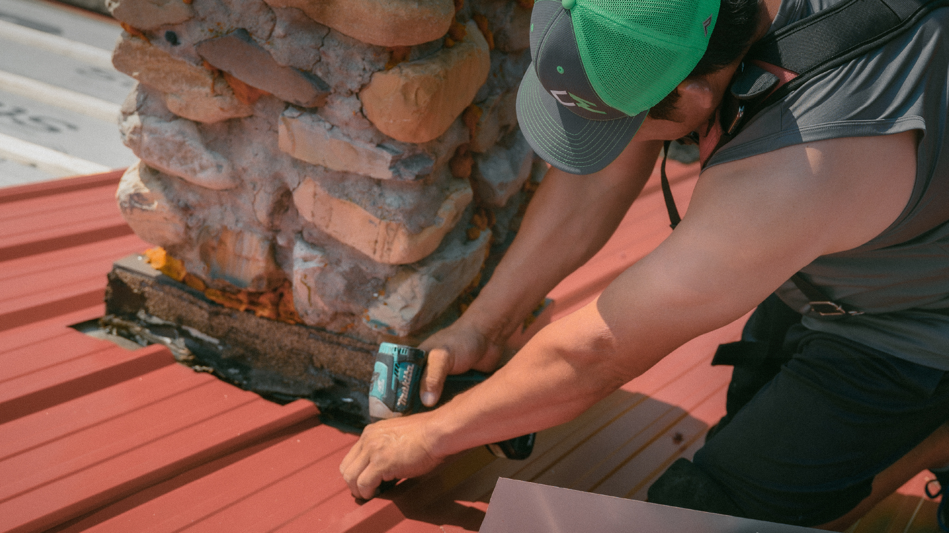 Close-up of a worker securing metal roofing over shingles near a chimney.