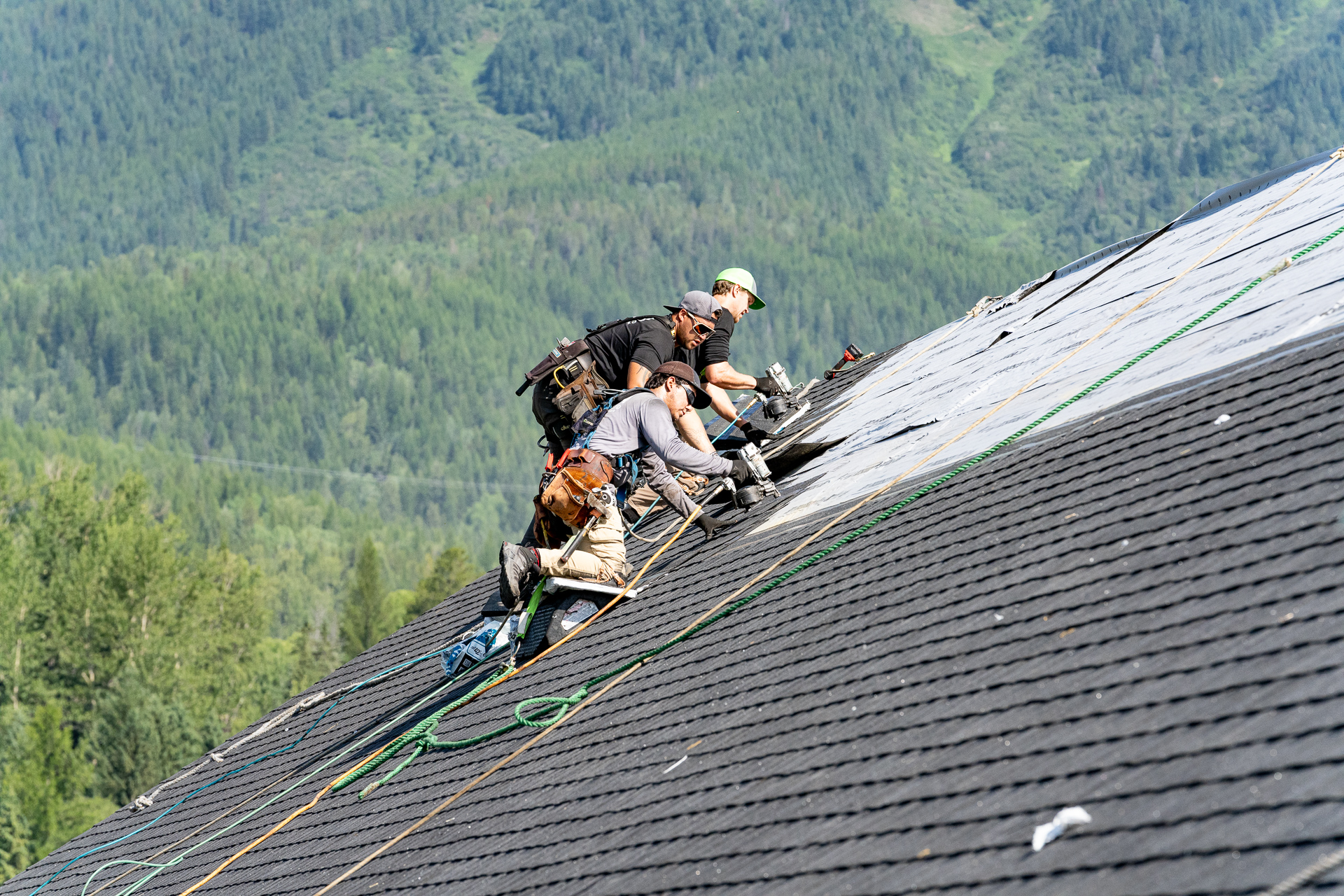 Roofers installing shingles on a steep roof with green forested hills in the background.