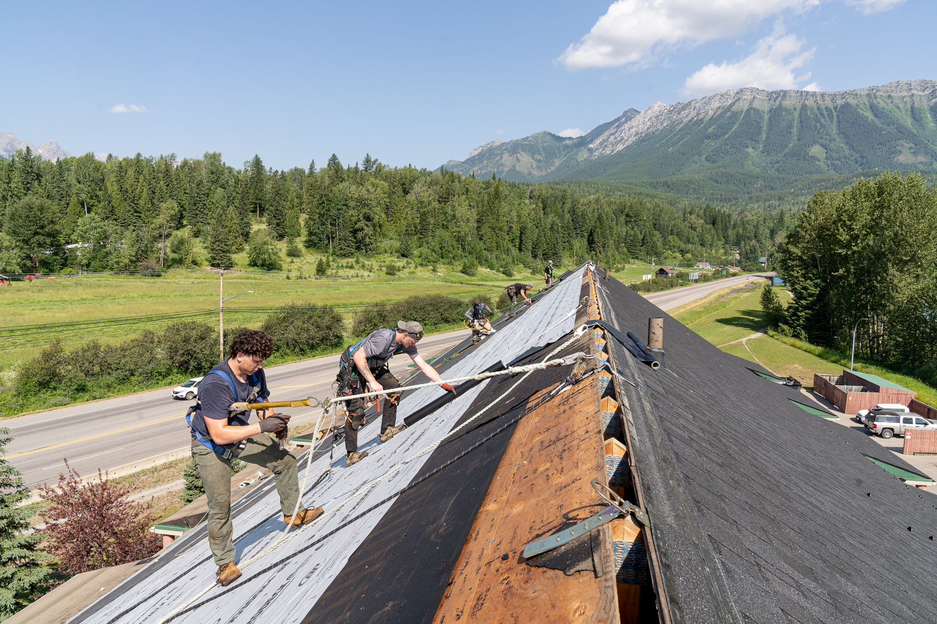 Roofers working on a large roof, installing underlayment with safety ropes, surrounded by green fields and mountainous terrain.