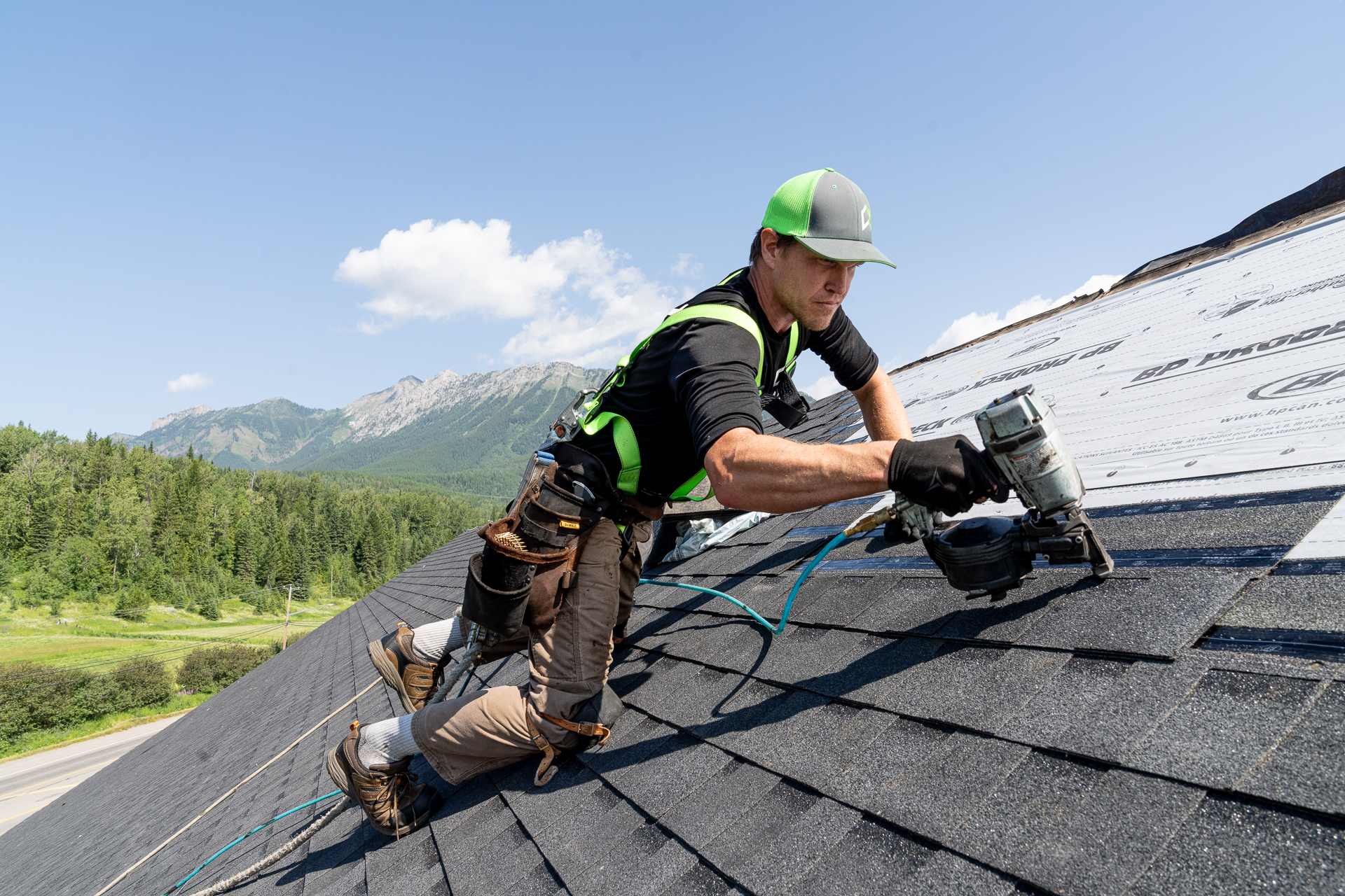 Roofer installing shingles on a steep roof with a scenic Cranbrook mountain background.