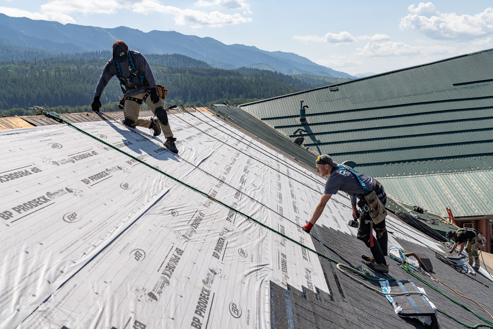 Two roofers working on a roof installation, adjusting the underlayment with safety lines attached, set against a mountain backdrop.