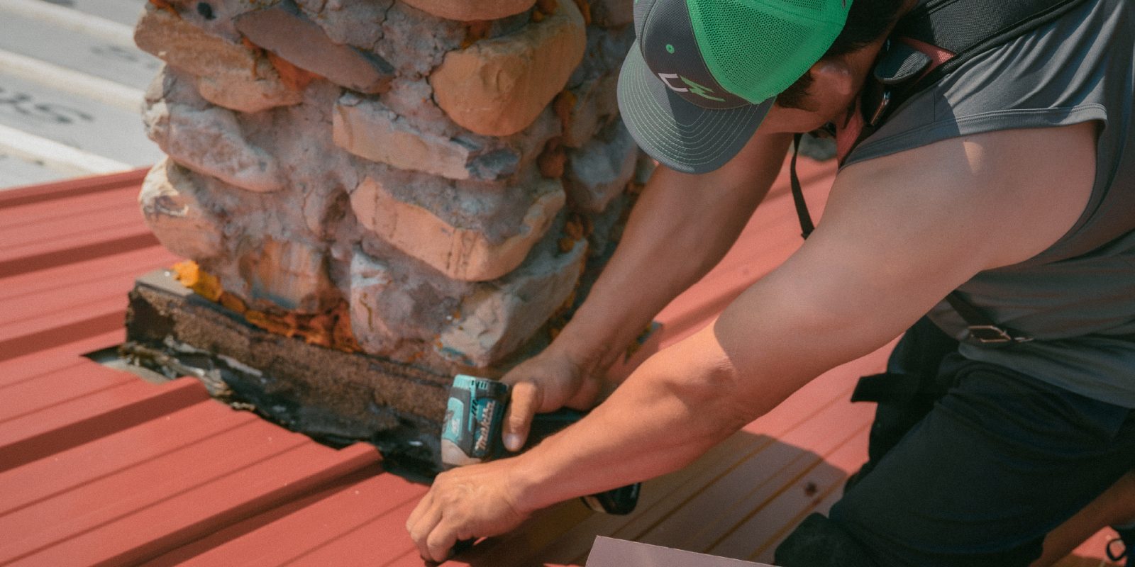 Close-up of a worker securing metal roofing over shingles near a chimney.