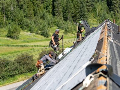 Three roofers installing roofing shingles on a steep roof with green fields and trees in the background.