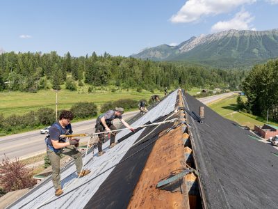 Roofers working on a large roof, installing underlayment with safety ropes, surrounded by green fields and mountainous terrain.