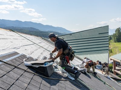 Roofers installing shingles on a steep roof with safety harnesses and equipment, with mountains visible in the background.