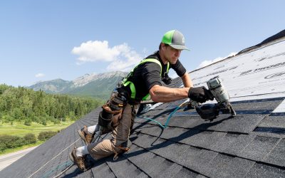 Roofer installing shingles on a steep roof with a scenic Cranbrook mountain background.