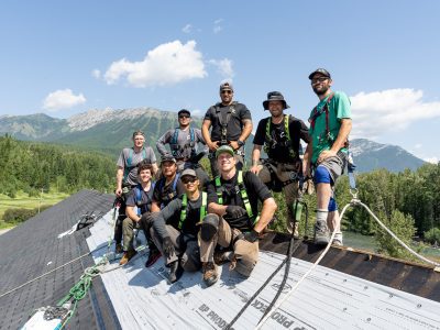 Group of roofers posing on top of a roof with harnesses, set against a Kootenay/Cranbrook mountain landscape.